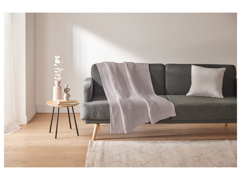 Een grijs-witte deken op een grijze bank, een beige vloerkleed en een houten bijzettafel.