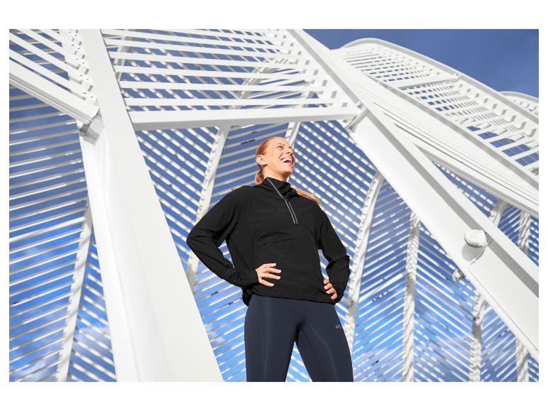 Vrouw in zwart sportjack en legging, lachend voor witte architectuur.