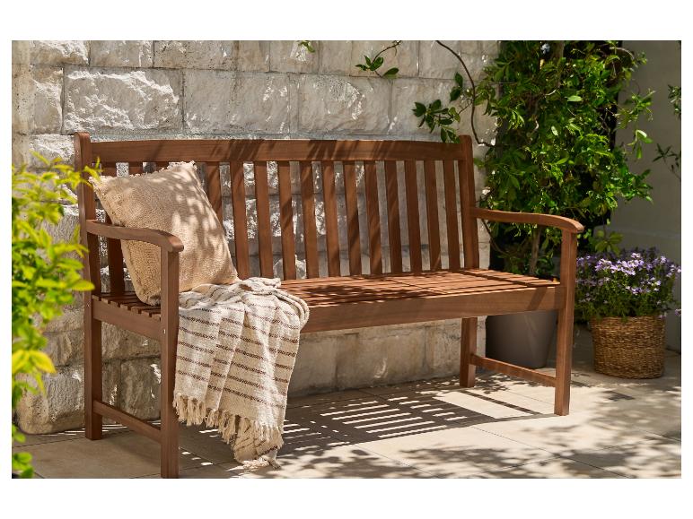 Houten tuinbank met een kussen en deken, tegen een stenen muur met planten.