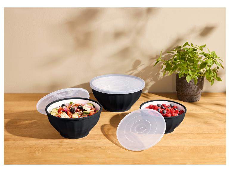 Drie zwarte kommen met transparante deksels, gevuld met salade en bessen, op een houten tafel.