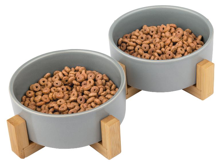 Twee grijze voerbakken voor huisdieren met houten standaards, gevuld met droogvoer.