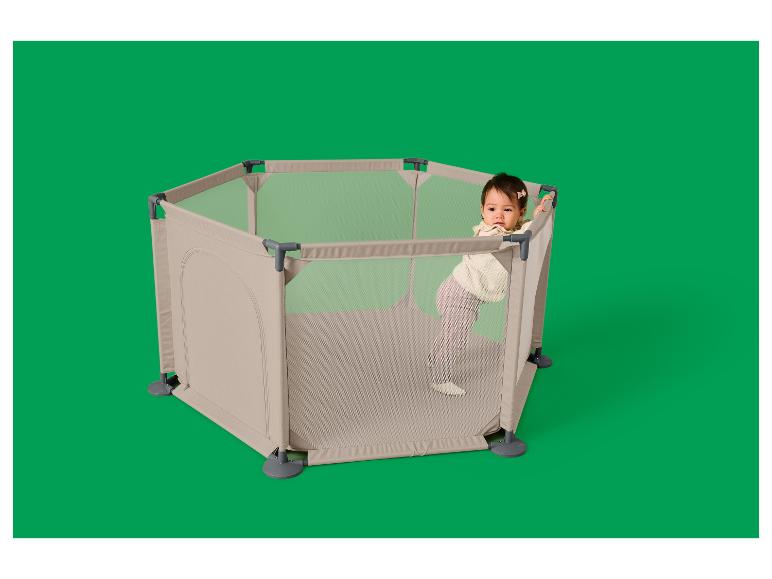 Baby in een zeshoekige, beige box met gaaswanden, staand en leunend op de rand.