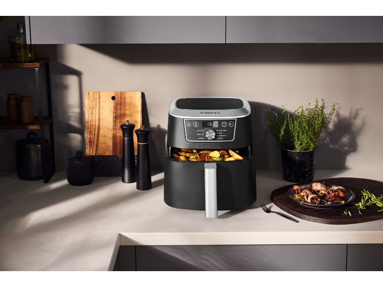 Zwarte airfryer met gebakken aardappelen in de keuken, naast kruidenmolens en een plant.