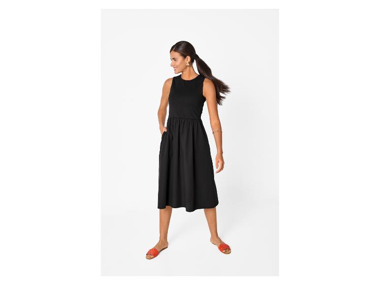 Vrouw in een zwarte mouwloze midi-jurk met zakken en oranje platte sandalen.