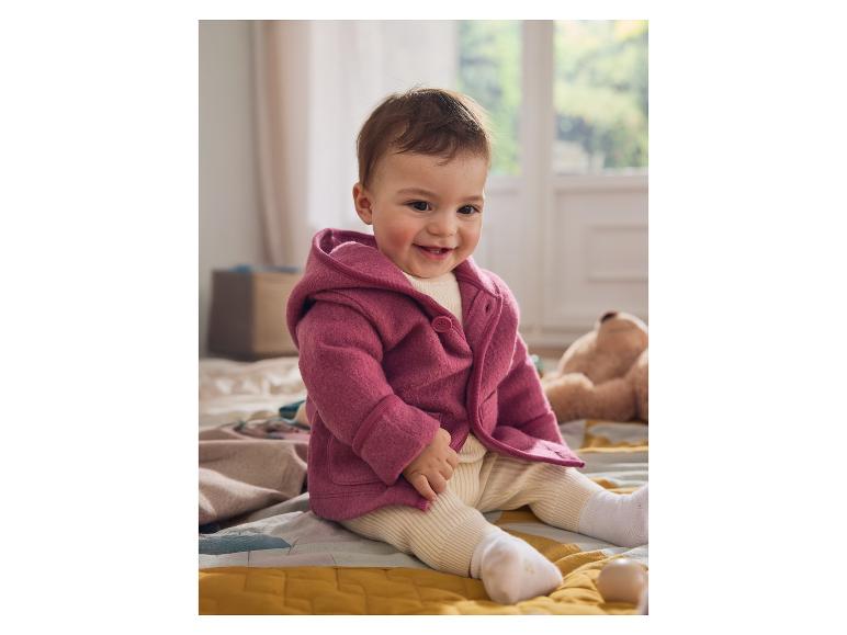 Een lachende baby in een roze jasje zit op bed.