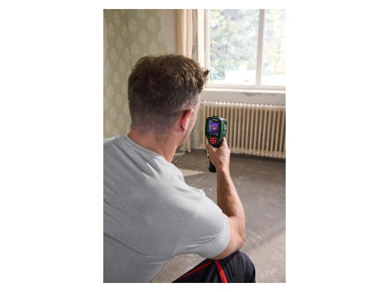 Man controleert kamertemperatuur met een warmtebeeldcamera in een kamer met oud behang en een radiator.