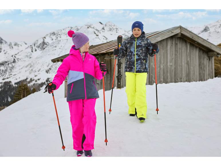 Twee kinderen in winterkleding met skistokken in een besneeuwd berglandschap met een houten hut.