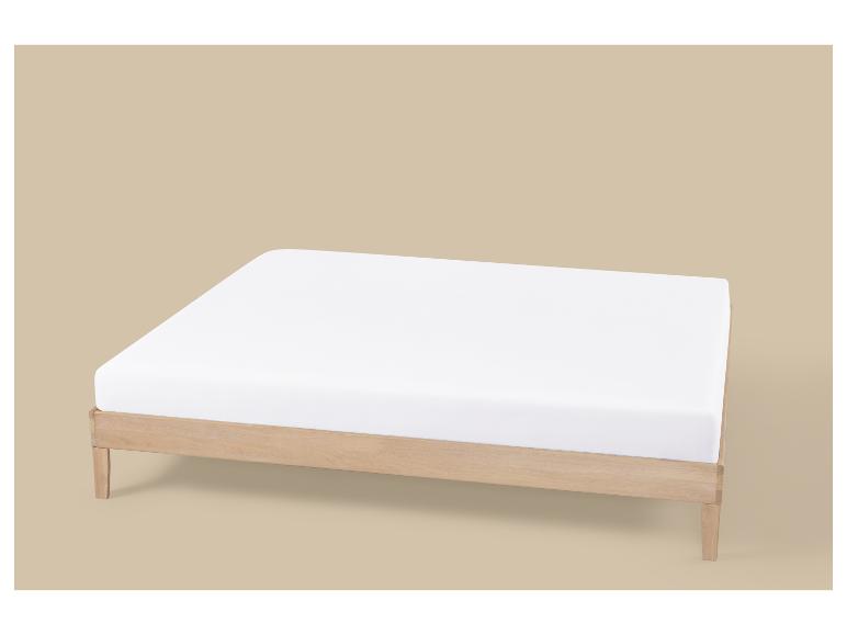 Houten bedframe met een wit matras op een lichtbruine achtergrond.