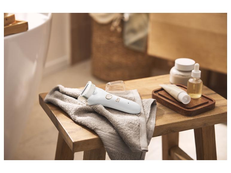 Vrouwenepilator op een handdoek, naast huidverzorgingsproducten op een houten krukje.