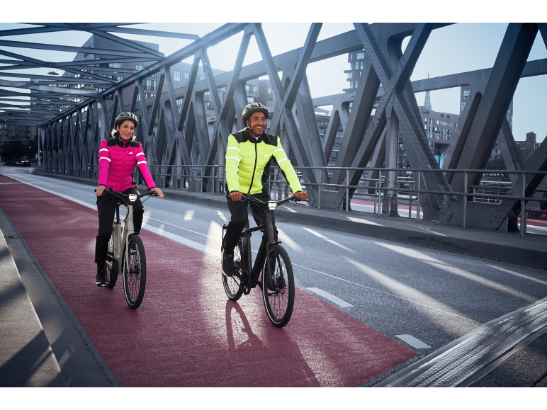 Twee mensen in reflecterende fietsjassen en helmen rijden op elektrische fietsen over een brug.
