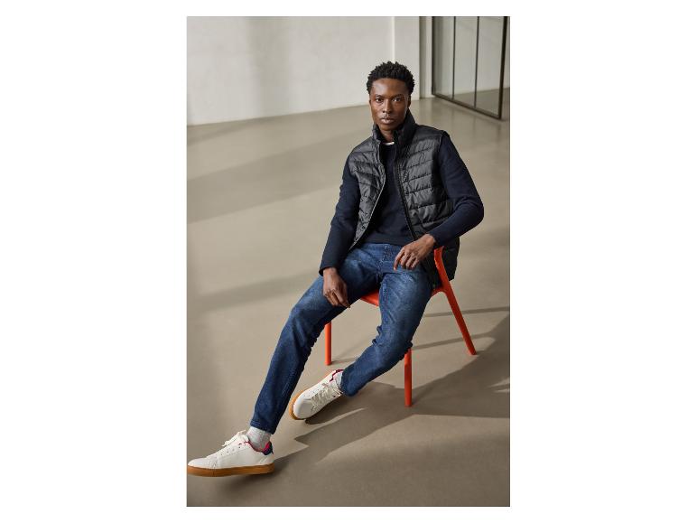 Man in zwart vest, marineblauwe trui en jeans, zittend op een oranje stoel.
