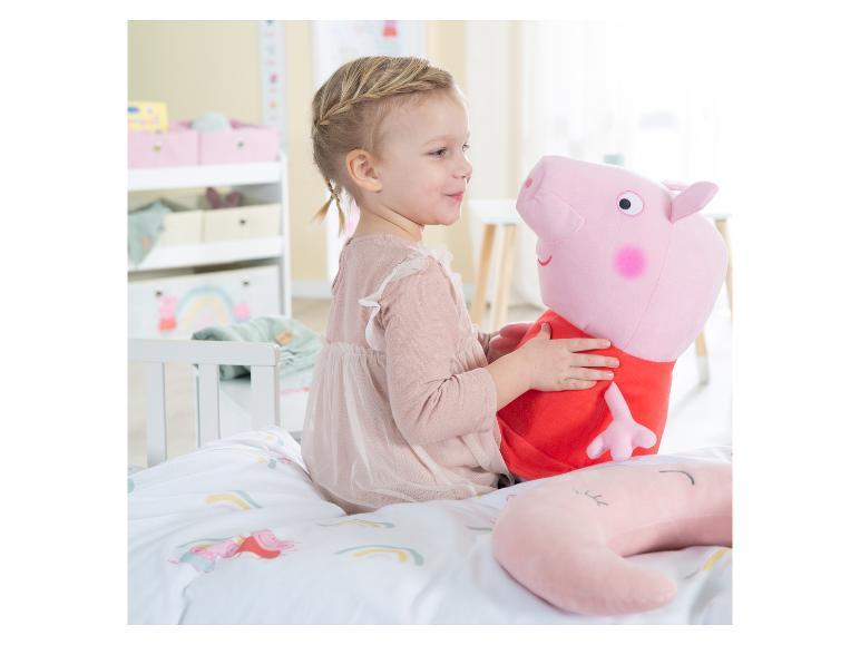 Een jong meisje in een roze jurk speelt met een Peppa Pig knuffel op een bed met een regenboogdekbedovertrek.
