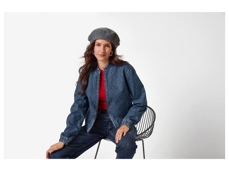 Vrouw in spijkerjack, rode top en baret, zittend op een stoel.