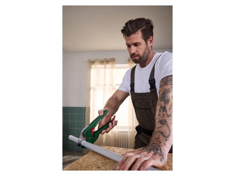 Een man snijdt een plastic buis met een groene PARKSIDE PVC-pijpsnijder op een houten tafel.