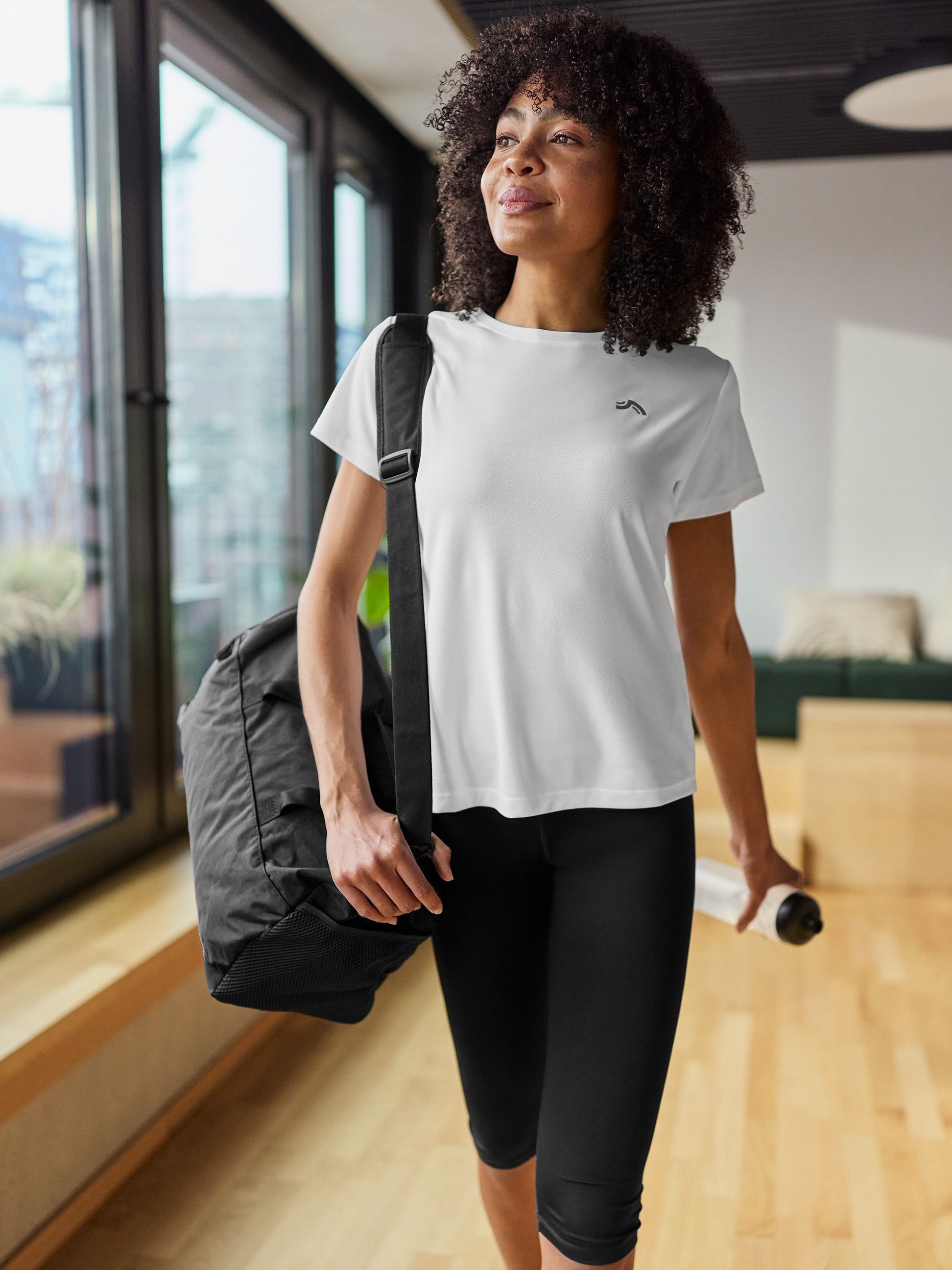 Vrouw in wit sportshirt, zwarte legging, met sporttas en waterfles.