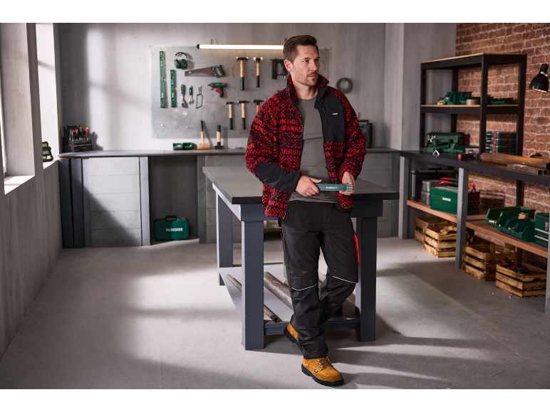 Man in rood-zwart fleecejack en werkbroek in een werkplaats.