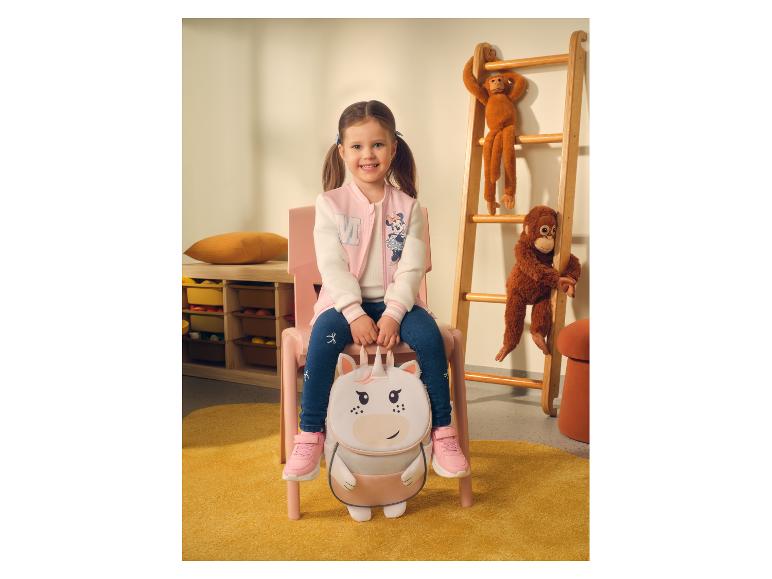 Meisje in baseballjack en jeans, zittend op een stoel met een eenhoornrugzak.