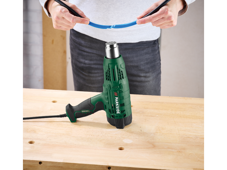 Groen heteluchtpistool op een houten werkbank, met een persoon die een blauwe kabel vasthoudt.