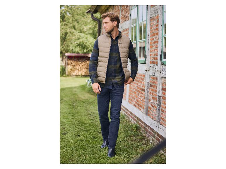 Man in een beige gewatteerd vest, geruit overhemd en jeans loopt buiten.