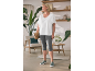 Vrouw in wit T-shirt en grijze capri legging, met gevlochten tas, staat in een gezellige kamer.
