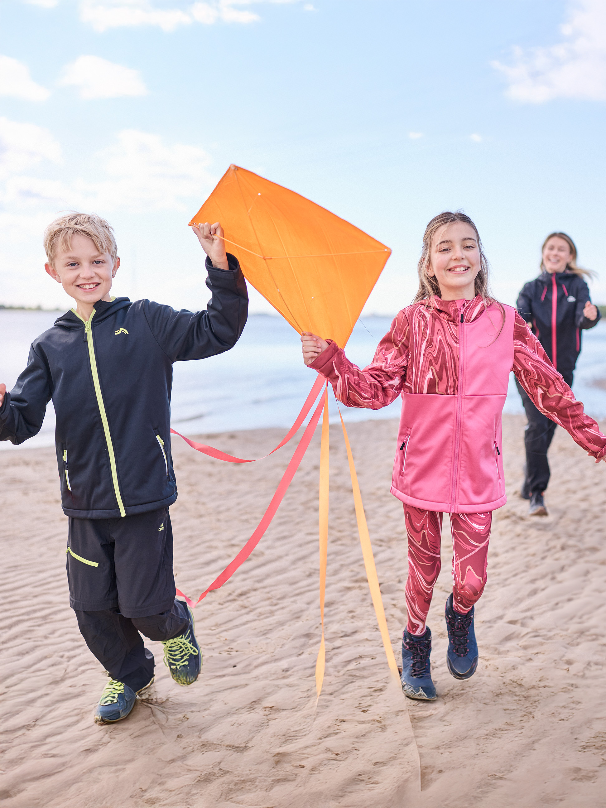 Kinderen in sportkleding rennen op strand met vlieger, vrouw op achtergrond.