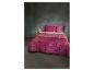 Roze beddengoed met patroon en kussens op een bed naast een wit bijzettafeltje en lamp.