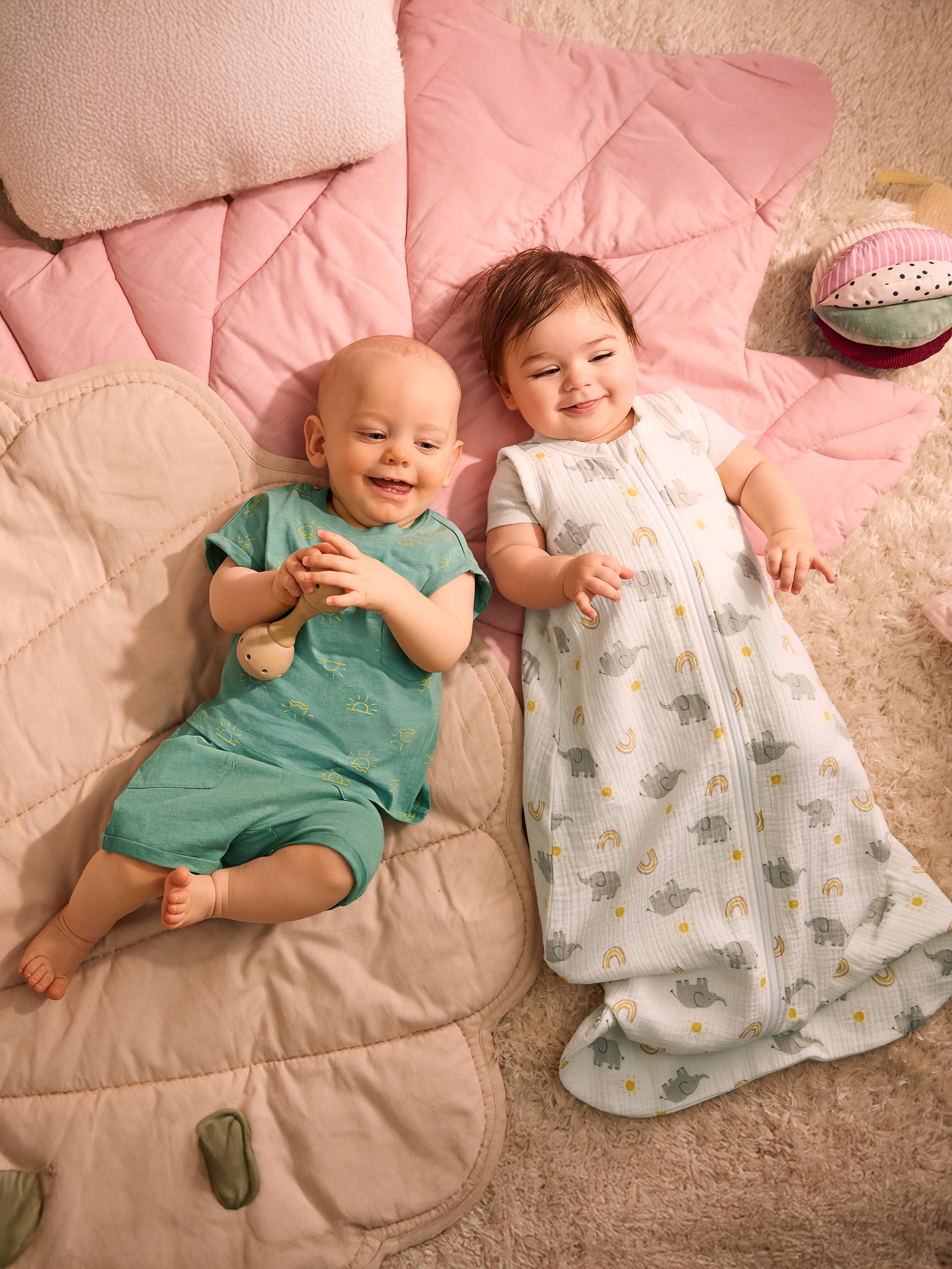 Twee baby's liggen op een roze deken, één in groene kleding, de ander in een slaapzak met olifanten.