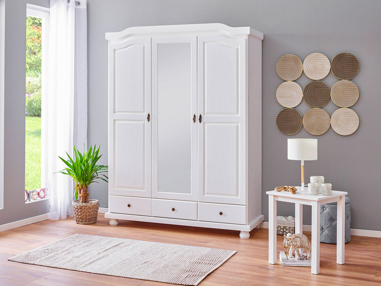 Een witte kledingkast met drie deuren en drie lades, naast een bijzettafel met een lamp.