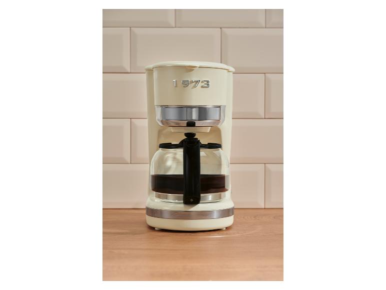 Crèmekleurig koffiezetapparaat met kan op houten aanrecht, tegen beige tegels.