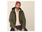 Man in een groene pufferjas, beige trui met halve rits, donkere jeans en een rode muts.