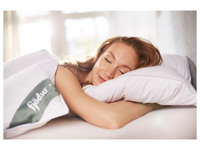 Vrouw slaapt op wit kussen en dekbed met Fjödur-logo in zonnige slaapkamer.
