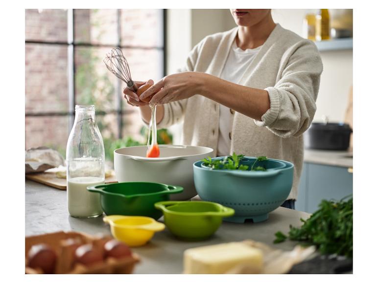 Een persoon breekt een ei in een kom met keukengerei en ingrediënten op tafel.