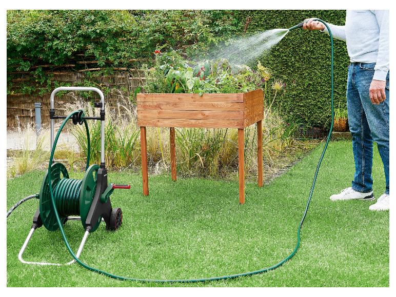 Man besproeit planten in een houten verhoogde moestuin met een groene tuinslang op een kar.