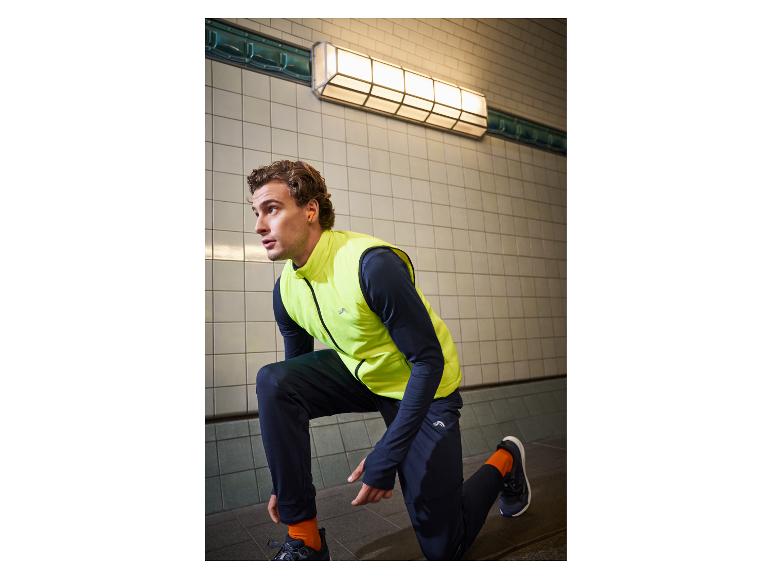 Man in geel sportvest en zwarte broek, gehurkt in een ondergrondse doorgang.