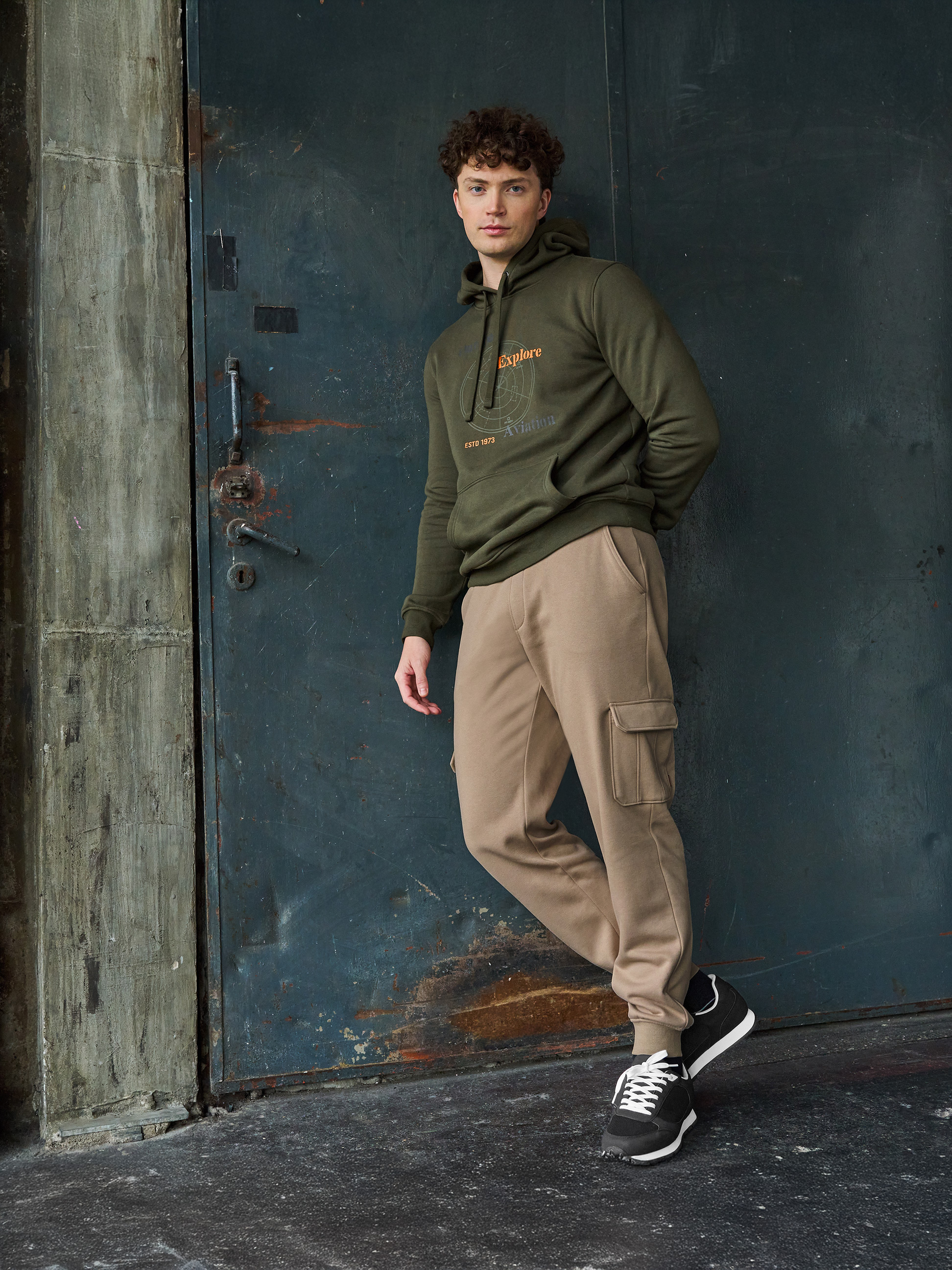 Man in olijfgroene 'Explore Aviation' hoodie en beige joggingbroek.