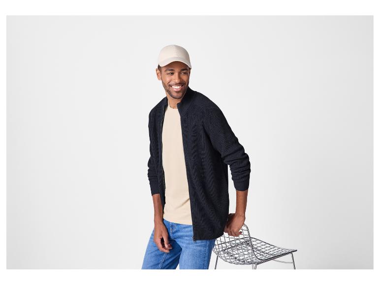 Lachende man met beige pet, marineblauwe cardigan en jeans.