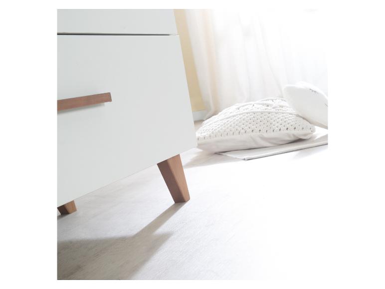 Witte ladekast met houten handgrepen en poten, naast een gebreid kussen op de vloer.