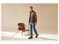 Man in bruin bodywarmer, geruit overhemd en jeans, naast een houten stoel.