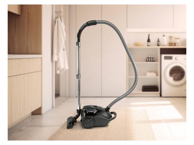 Zwarte stofzuiger op een vloerkleed in een wasruimte met houten kasten en een wasmachine.