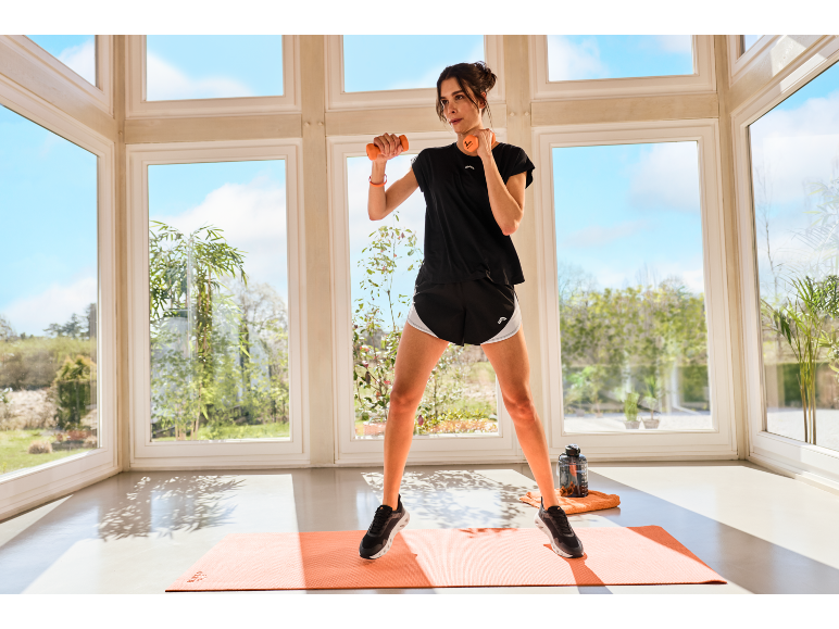Vrouw in sportkleding traint met dumbbells op een oranje mat.