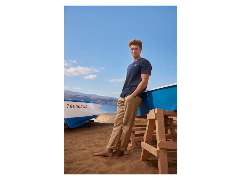 Man in een marineblauw T-shirt en beige broek op het strand, leunend tegen een boot.
