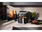 Zwarte airfryer met gebakken aardappelen in de keuken, naast kruidenmolens en een plant.