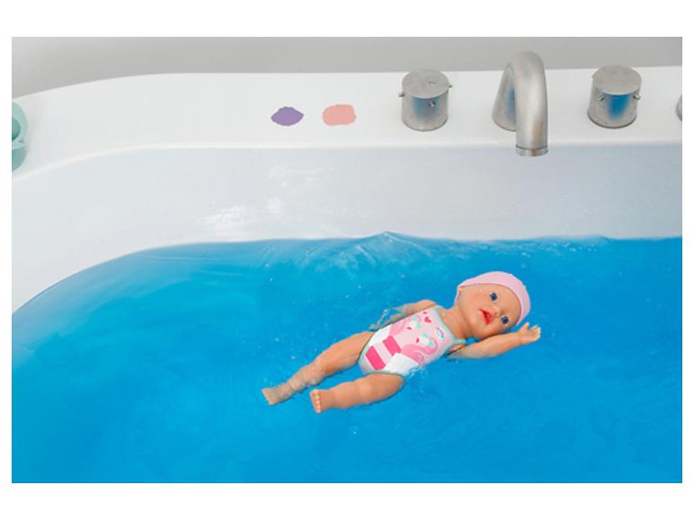 Babypop zwemt in blauw water in een bad, gekleed in een roze zwempak.