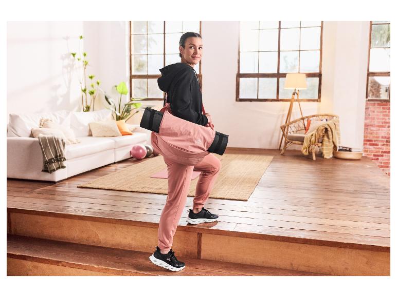 Vrouw in roze broek en zwarte trui loopt de trap op met een Crivit yogatas.