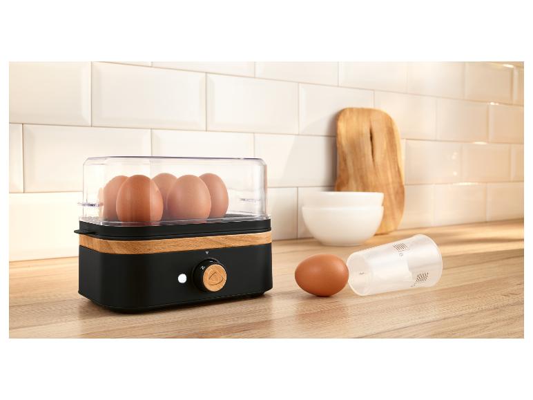 Eierkoker met houten accenten, eieren en maatbeker op een aanrecht.