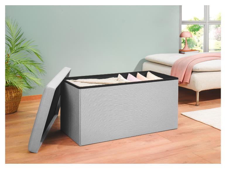 Een grijze opbergkist van stof met een open deksel, waar kussens en dekens in te zien zijn, staat op een lichte houten vloer.