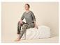 Vrouw in pyjama met paddenstoelprint zit op een witte poef, kijkt omhoog en lacht.