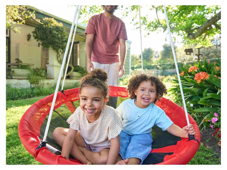Twee lachende kinderen in een rode tuinschommel, met vader op de achtergrond.
