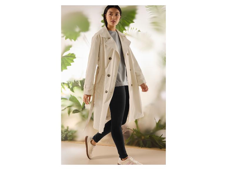 Vrouw in beige trenchcoat, lichtgrijze top, zwarte legging en witte sneakers.