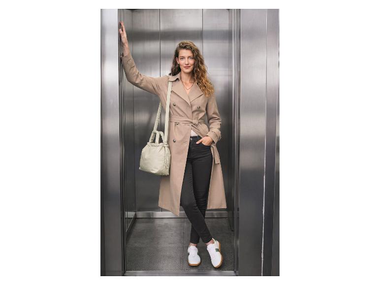 Vrouw in trenchcoat, zwarte jeans en witte sneakers, met een lichtgroene tas in een lift.